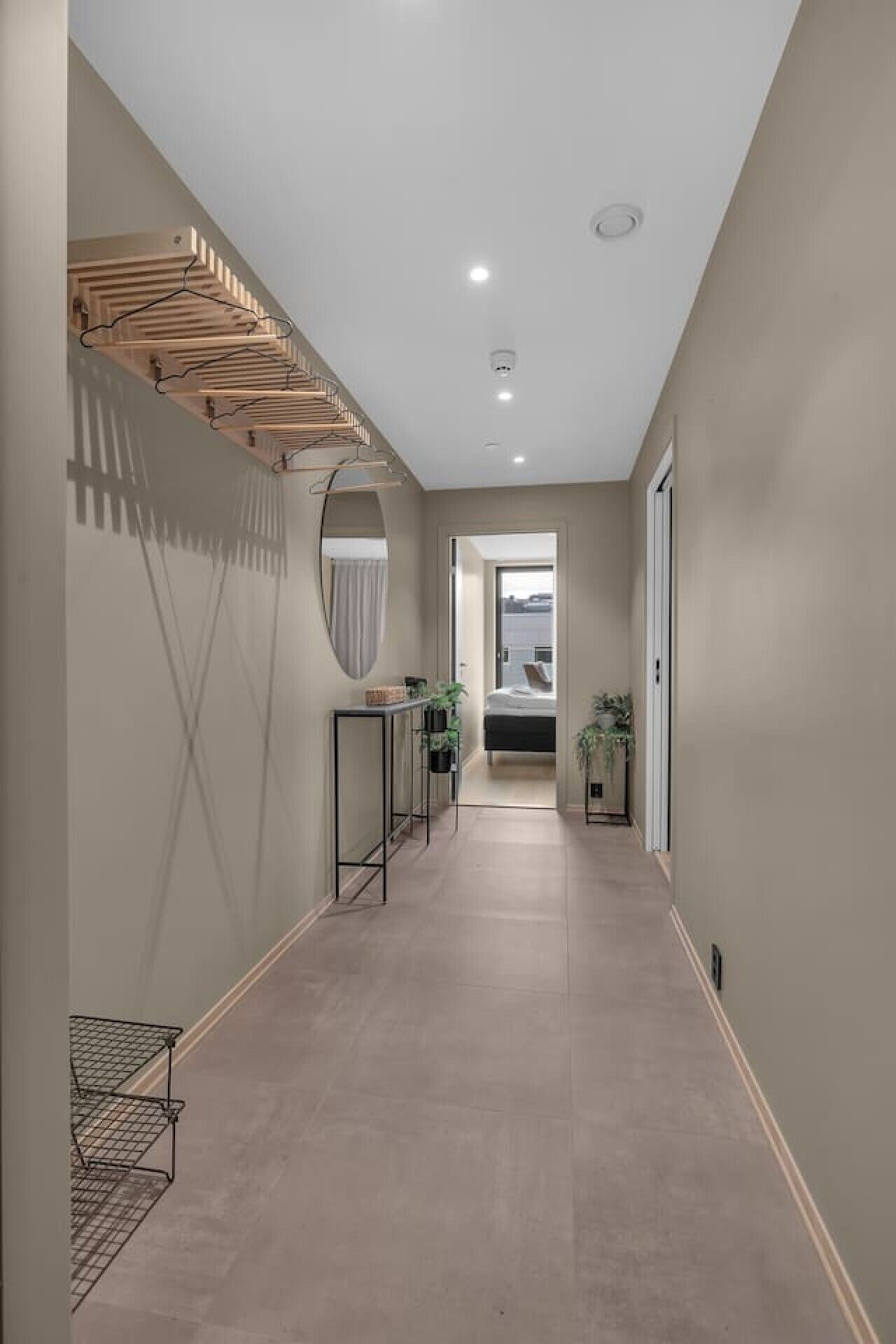 A hallway with a wooden drying rack, a mirror, a small table with plants, and a bed in the distance.