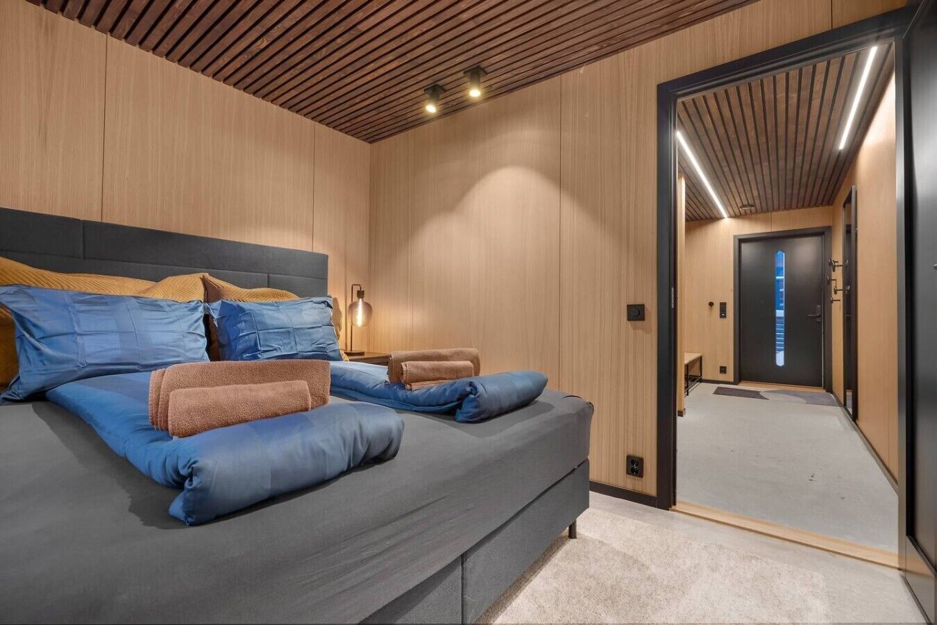 A modern bedroom with a large bed, wooden panel walls, and a mirror reflecting the room.
