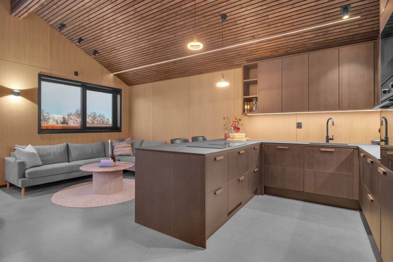 A modern kitchen with wooden cabinets, a built-in island, and a grey sofa in a living area with a large window.