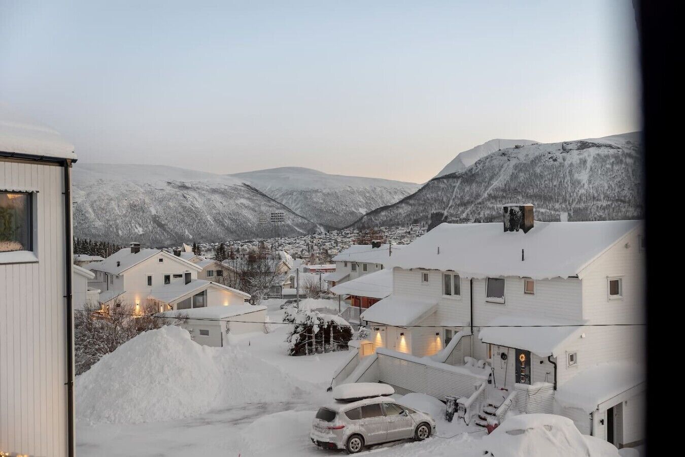 A residential area with houses covered in snow, a car parked in front, and mountains in the background.