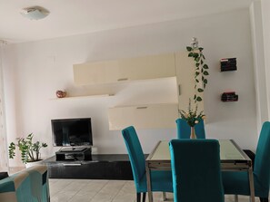 Una sala de estar moderna con un televisor de pantalla plana, sillas color turquesa, una mesa de comedor con cubierta de vidrio y un diseño de pared minimalista.
