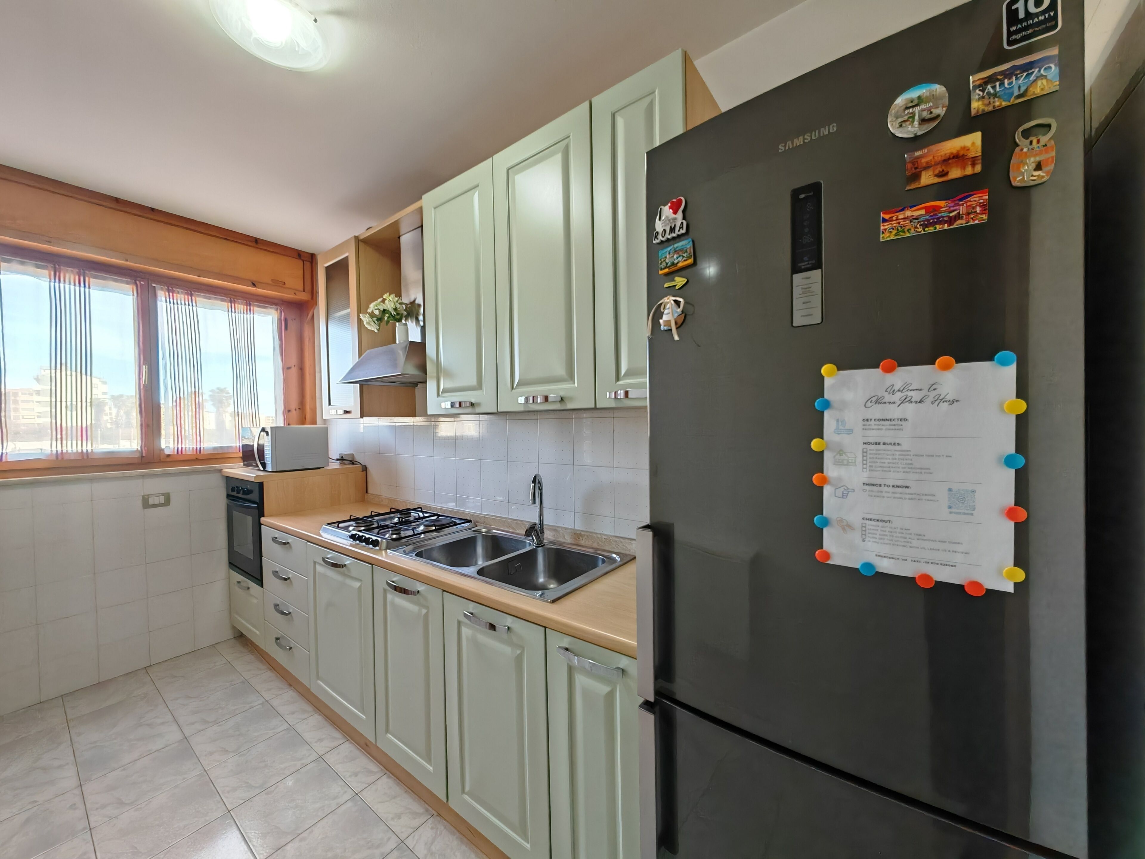 A kitchen with a stainless steel refrigerator, a microwave, a gas stove, and a sink. There are light green cabinets and a window with curtains.