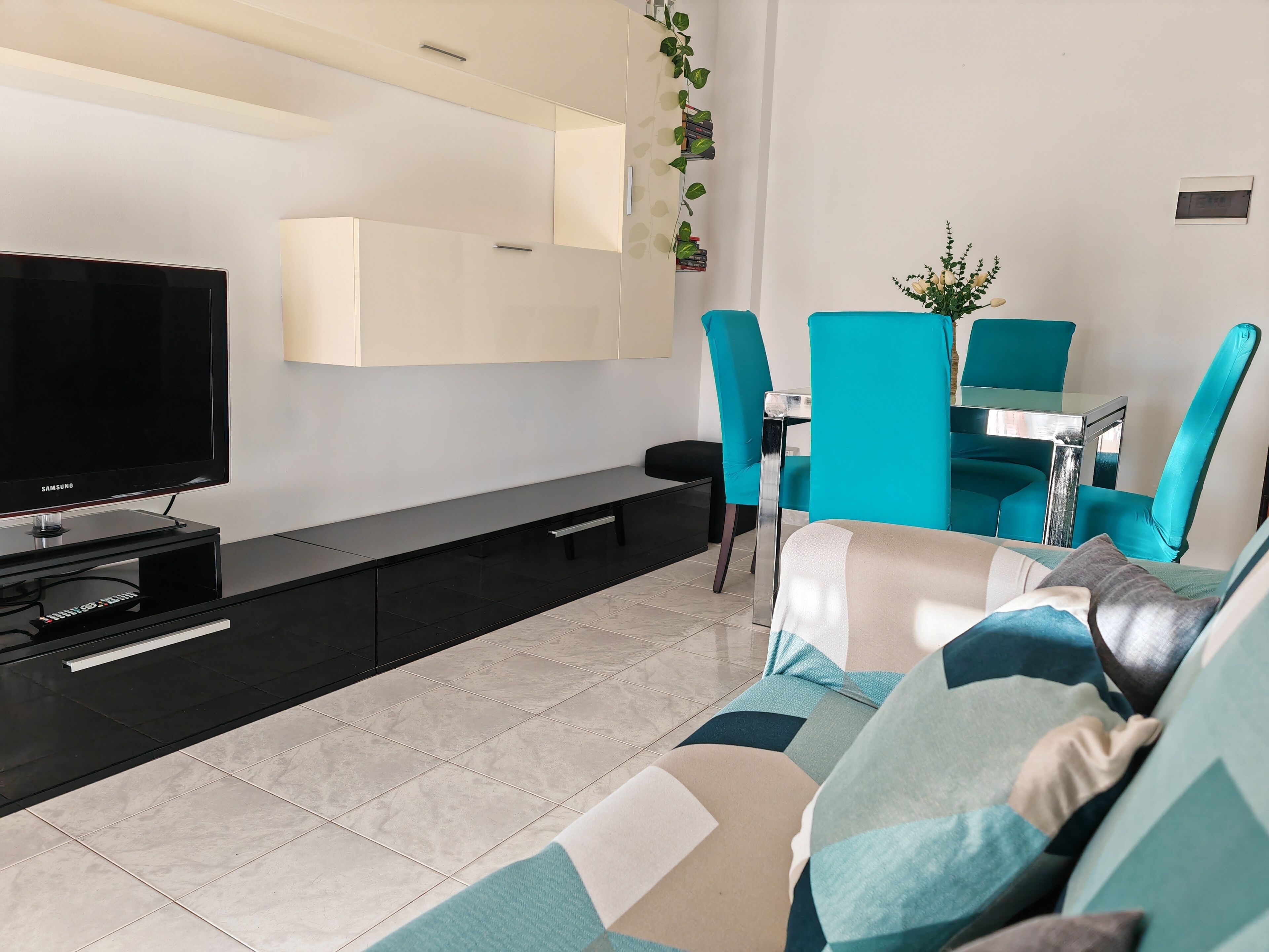 Un moderno salón con televisión de pantalla plana, un sofá, una mesa de centro y un comedor con sillas color turquesa.