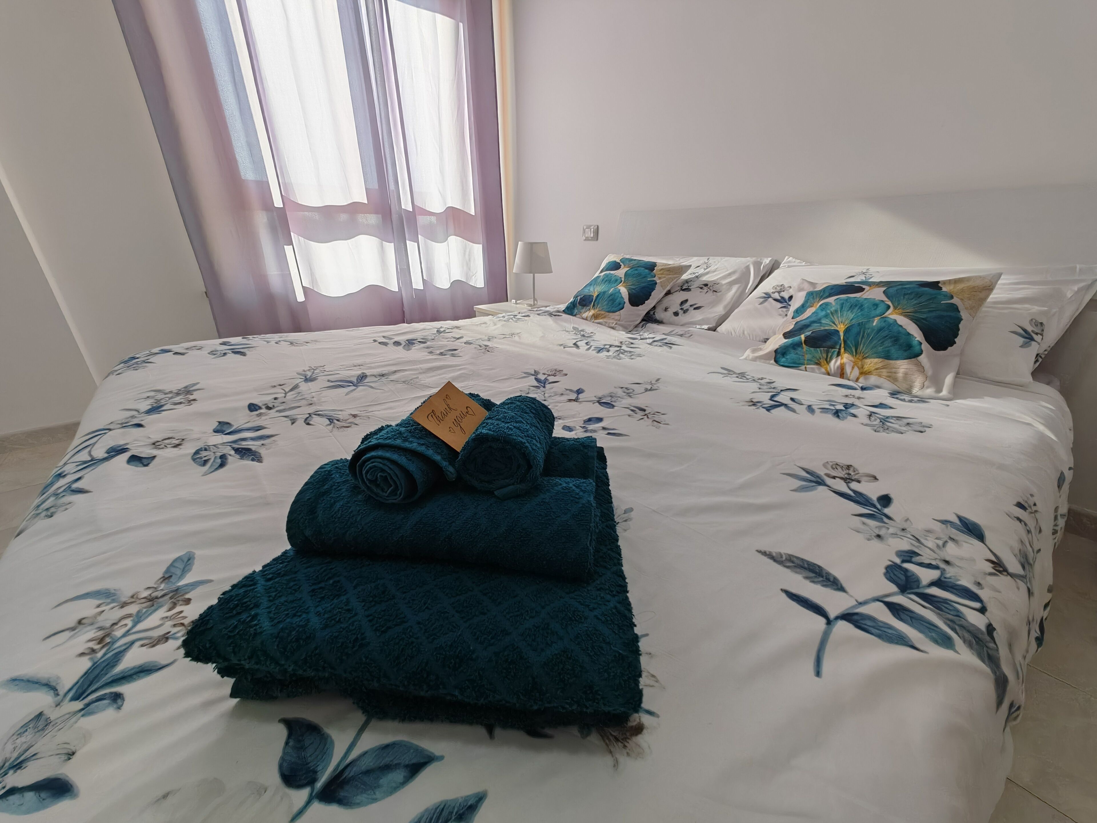Una cama bien hecha con ropa de cama floral, una nota y toallas enrolladas.