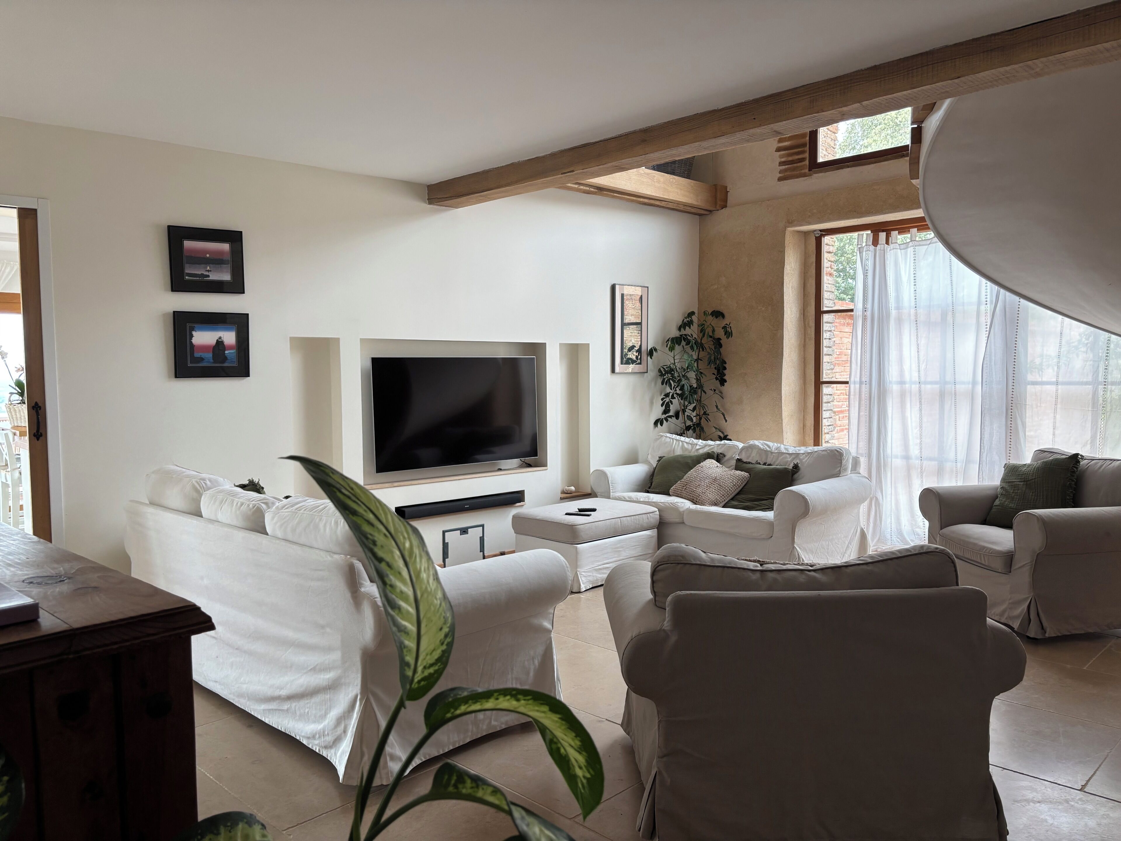 Een woonkamer met een flatscreen televisie, witte banken, een salontafel en een grote plant.