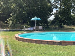 A circular swimming pool with a surrounding grassy area, a table with chairs, and a blue umbrella.