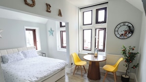 Ein Schlafzimmer mit einem Bett, einem runden Holztisch, zwei gelben Stühlen, einem Blumentopf, einer Wanduhr und einer Stern-Dekoration.