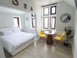 A bedroom with a bed, a round wooden table, two yellow chairs, a potted plant, a wall clock, and a star decoration. - The Ocean House - Sea Whisper (Hunstanton)