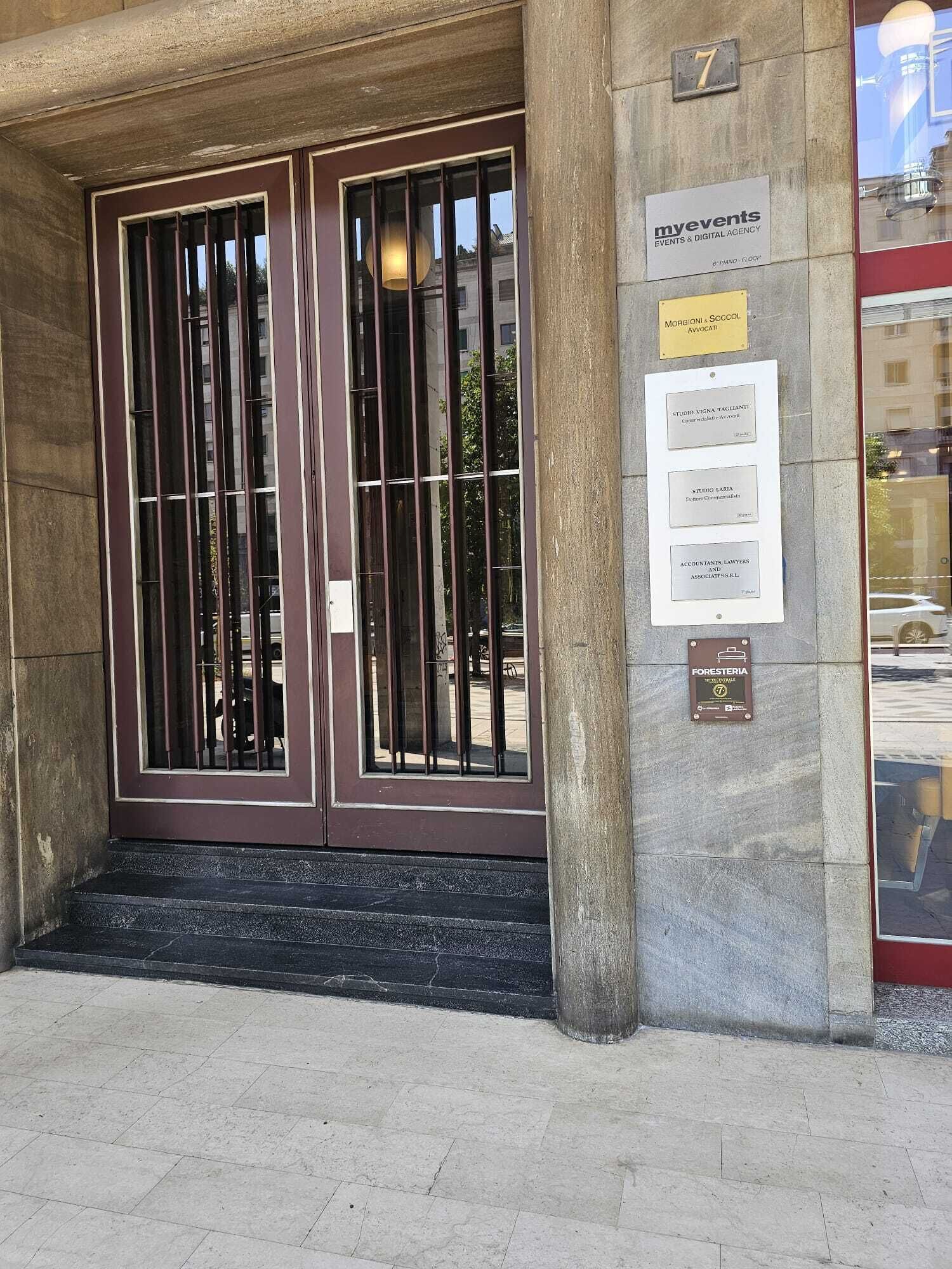 Een gebouwde ingang met een dubbele deur, een bord met 'myevents EVENTS & DIGITAL AGENCY' en plaquettes voor 'MORGIONI & SOCCOL AVVOCATI' en 'FORESTERIA'.