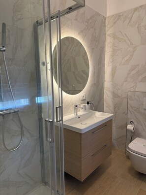 Een moderne badkamer met een douchecabine van glas, een ronde spiegel, een witte wastafel en een toilet.