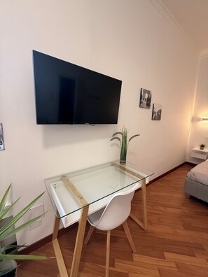 Een moderne woonkamer met een flatscreen televisie aan de muur, een salontafel met glazen blad, een witte fauteuil, een kleine potplant en een bed met witte dekbedovertrek.
