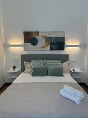 Een net opgemaakt bed met een geborduurde spreidek, kussens en een opgerolde handdoek. Twee nachtkastjes met een plant en een afstandsbediening.