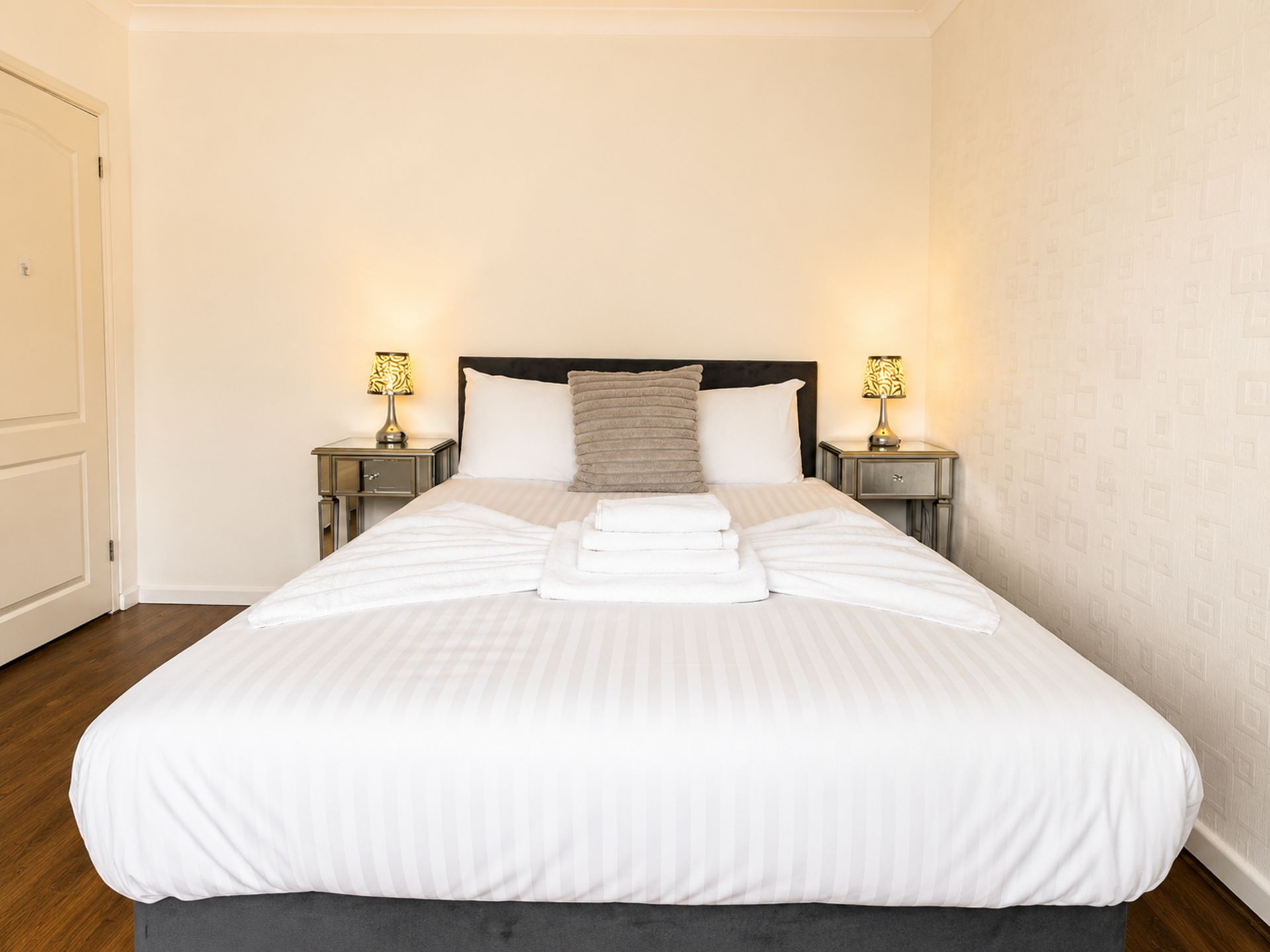 Una cama bien hecha con sábanas blancas y un cabecero oscuro, flanqueada por dos mesitas de noche con lámparas, en una habitación con paredes lisas y suelo de madera.