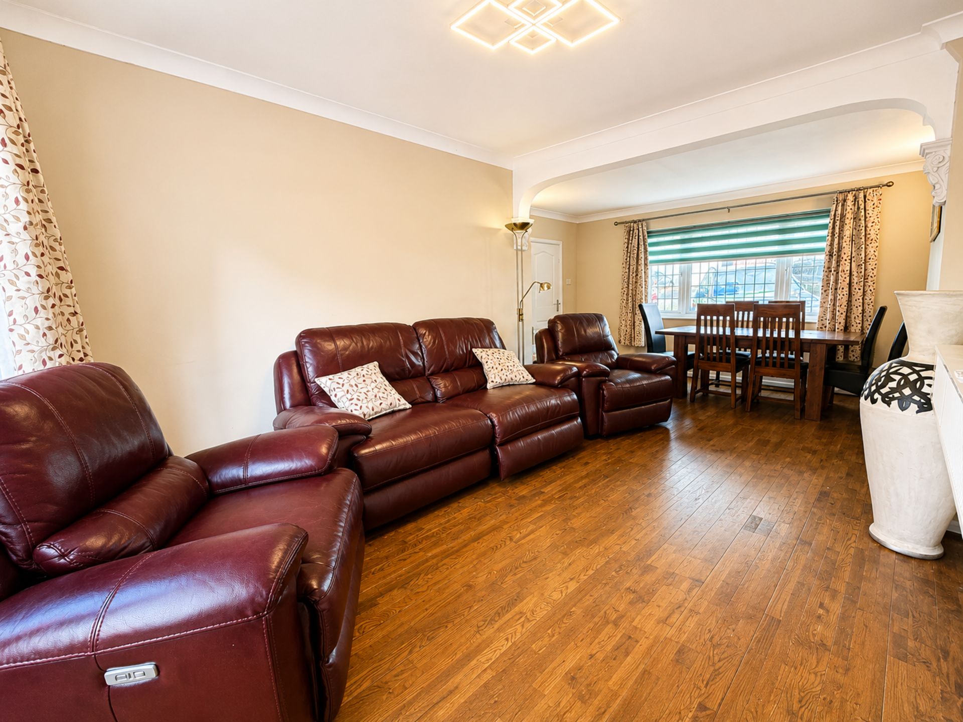 A living room with a brown leather sofa, a dining table with chairs, and a large window with curtains.