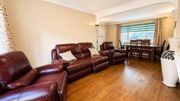 A living room with a brown leather sofa, a dining table with chairs, and a large window with curtains.