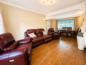 A living room with a brown leather sofa, a dining table with chairs, and a large window with curtains.
