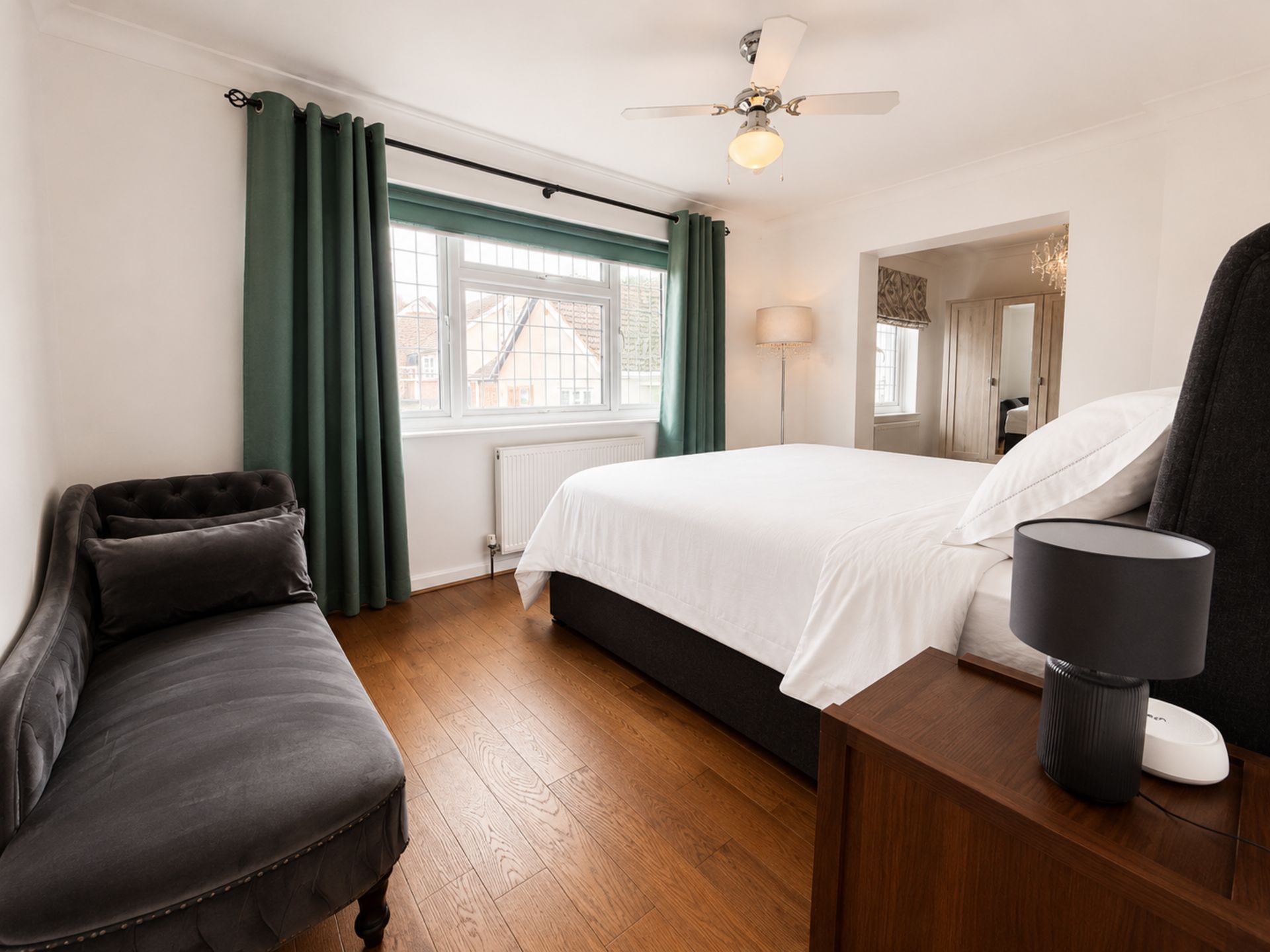 Et hotelværelse med seng, skrivebord, stol, gulvlampe, ventilator i loftet og et vindue med gardiner.