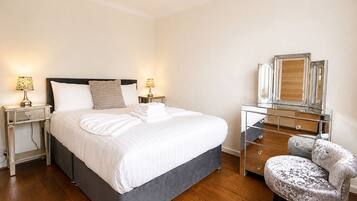 A bedroom with a large bed, two bedside tables with lamps, a dresser with drawers, a chair, and a mirror.