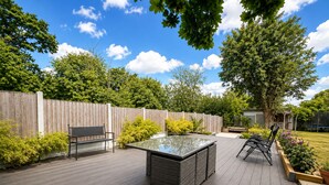 A wooden deck with a glass-top table and chairs, surrounded by a wooden fence and greenery. - Spacious 4BDR Home in Rayleigh Essex (Essex)