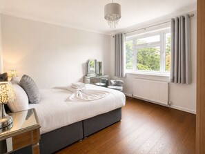 Quarto com cama, mesa de cabeceira, abajur, cômoda e janela com cortinas.