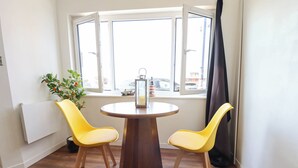 A room with a round wooden table, two yellow chairs, a potted plant, and a window with curtains. - The Ocean House - Sea Shell (Hunstanton)