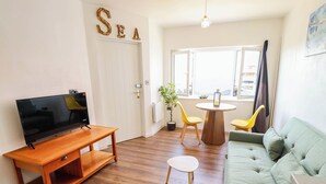A living room with a television, wooden furniture, a green sofa, and a dining area with yellow chairs. - The Ocean House - Sea Shell (Hunstanton)