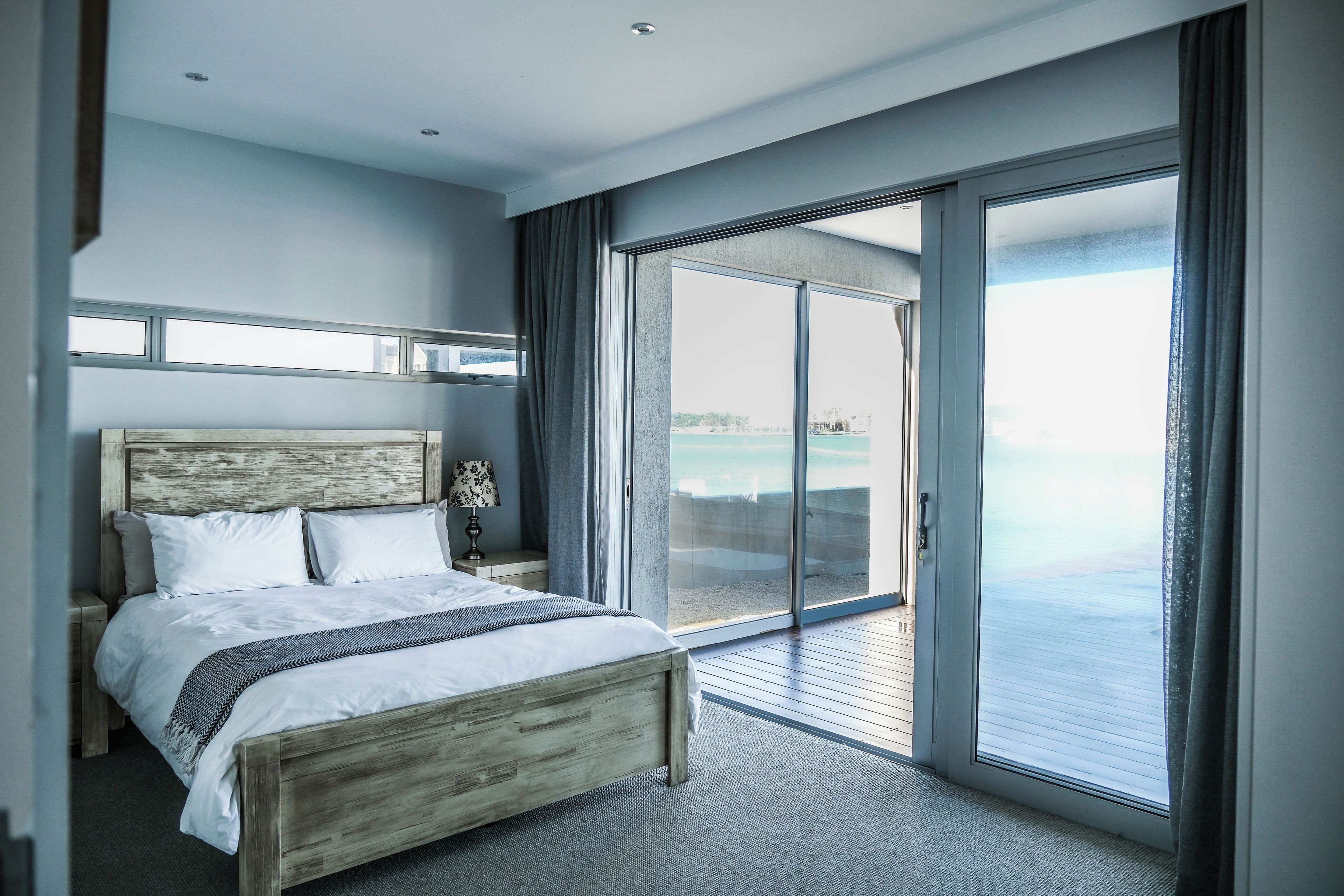 A modern bedroom with a large bed, a nightstand, a lamp, and a view of the sea through a sliding glass door.