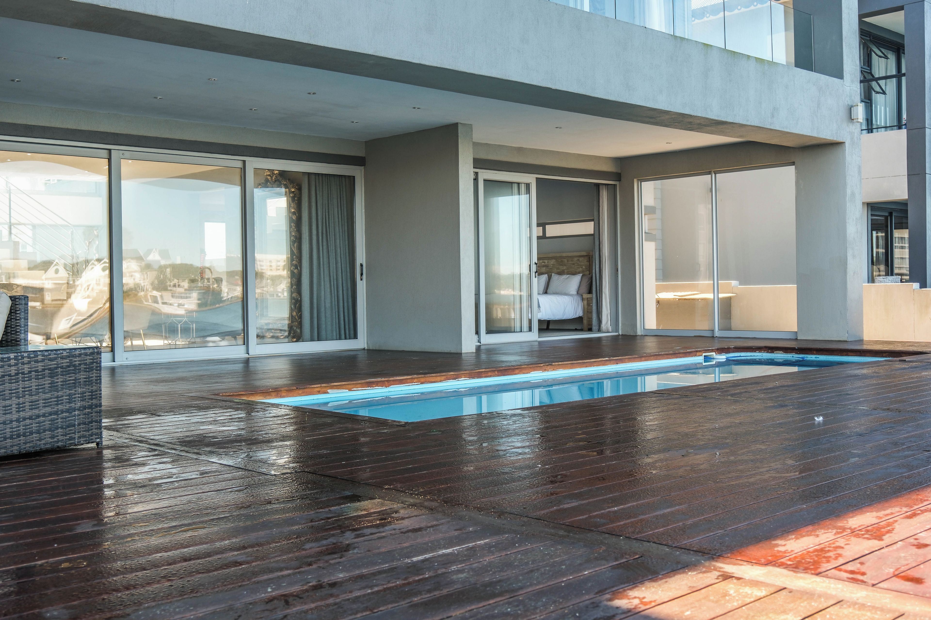 A modern outdoor swimming pool area with a wooden deck, glass doors leading to a bedroom, and a view of the cityscape.