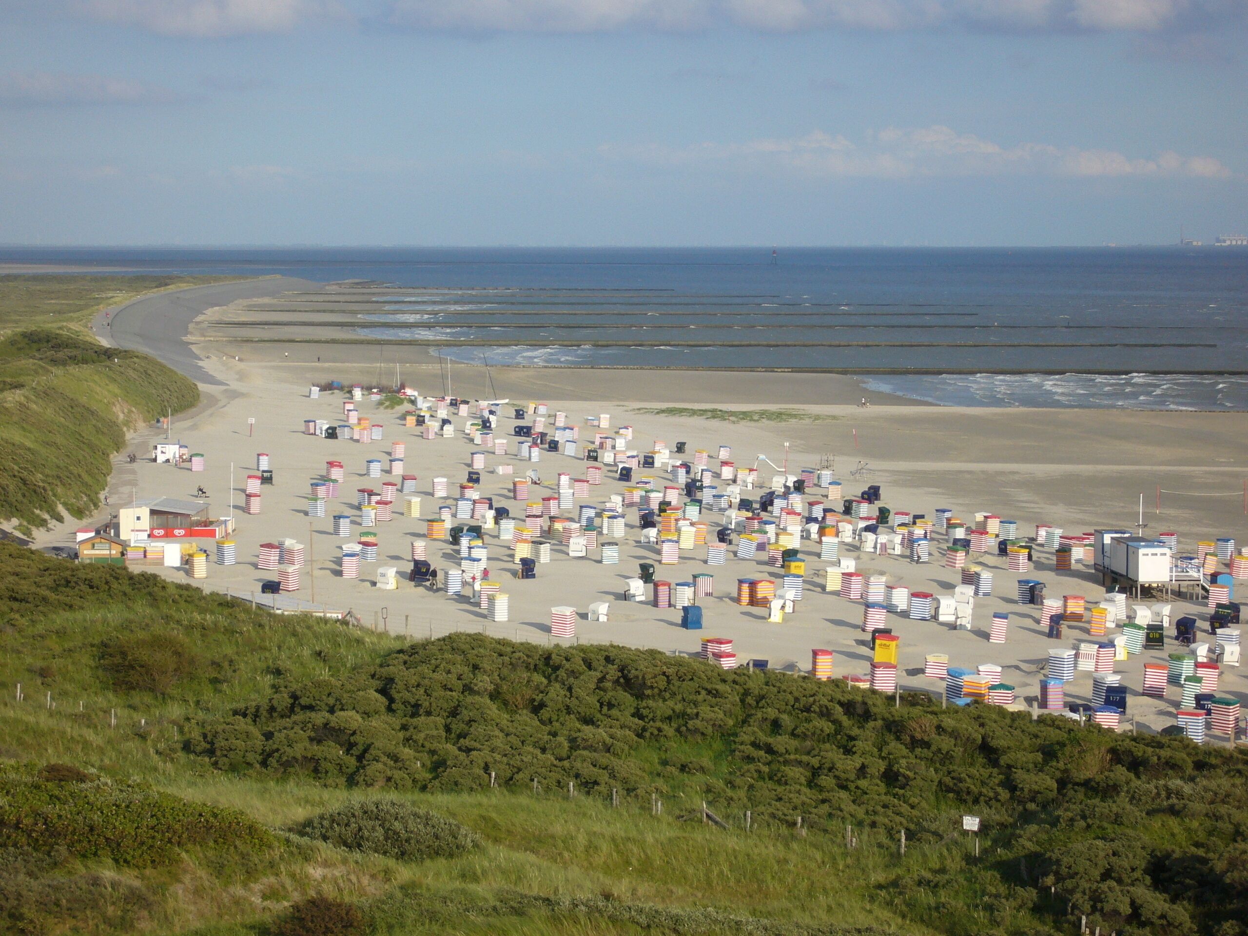 Ein Strand mit zahlreichen bunten Behältern, die im Sand gestapelt sind, eine sanfte Hügelkuppe im Vordergrund und ein klarer Himmel.
