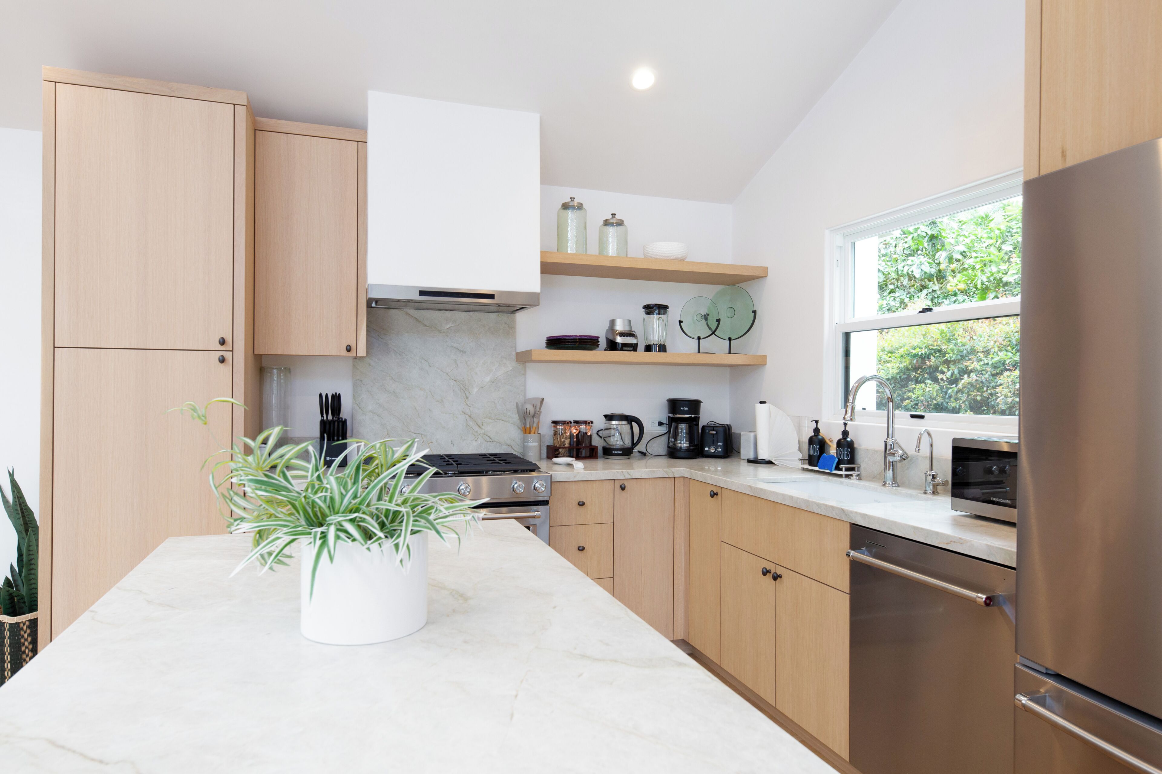 Een moderne keuken met houten kastjes, een marmeren aanrechtblad, een roestvrijstalen koelkast en een raam met uitzicht op groen.