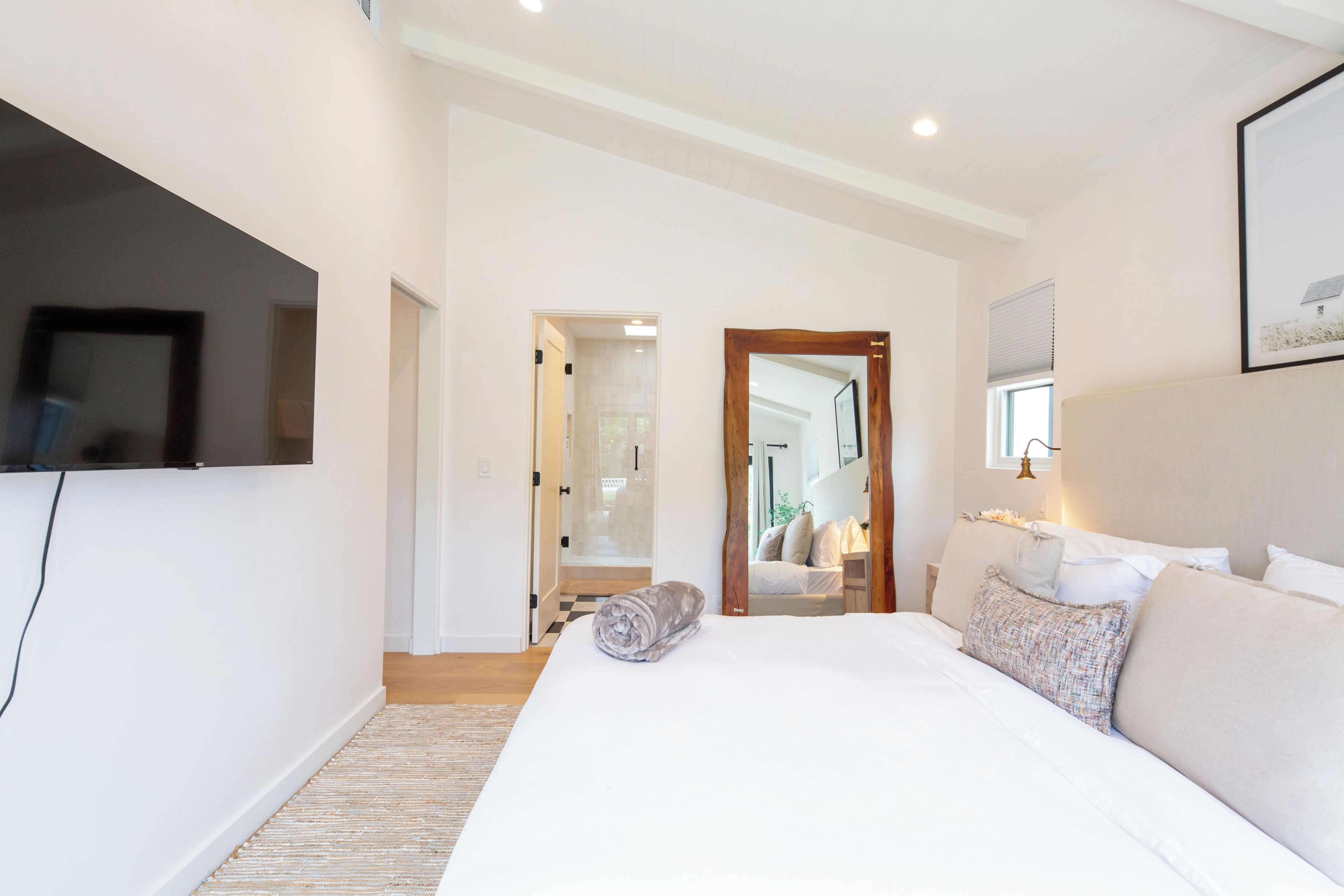 A bedroom with a large bed, a flat-screen TV mounted on the wall, and a mirror reflecting the room.