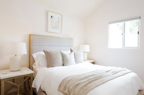 Een slaapkamer met een bed, nachtkastjes, lampen, een raam en een ingelijst schilderij aan de muur.