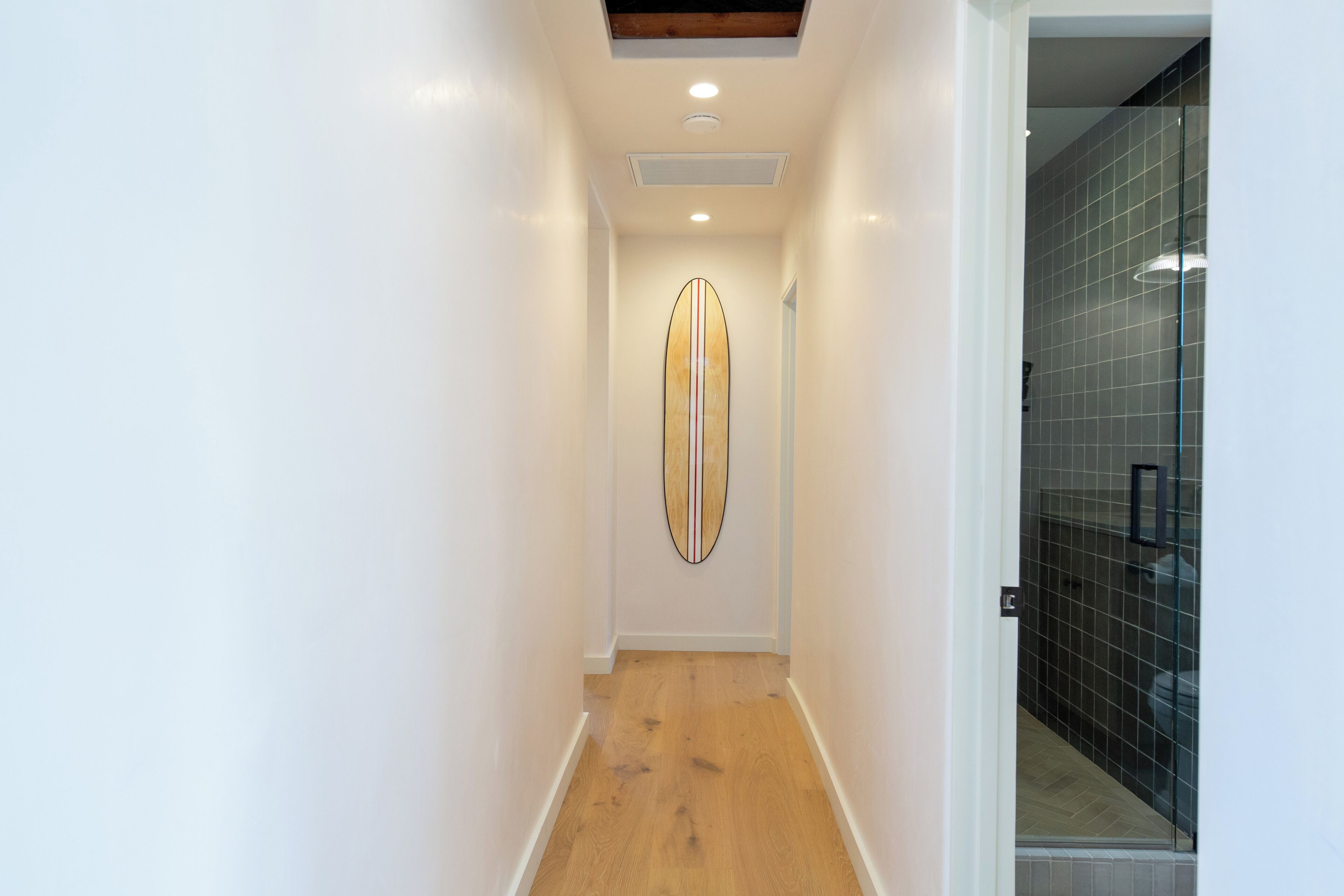 A narrow hallway with a wooden floor, a ceiling with recessed lighting, and a surfboard mounted on the wall.