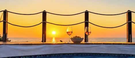 Une piscine avec vue sur le coucher de soleil, une table à manger avec des verres et un saladier, et une barrière faite de cordes.