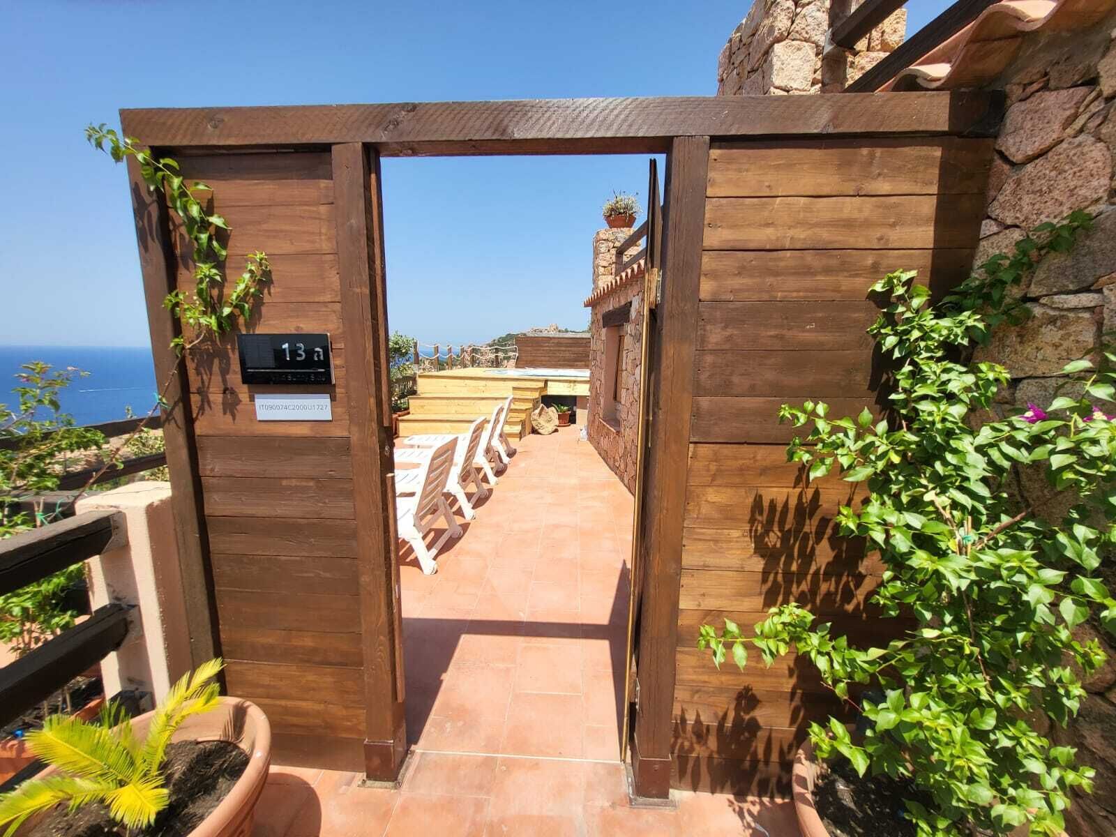 A wooden gate leading to a terrace with a stone wall, a sign with the number 13, and a view of the sea.