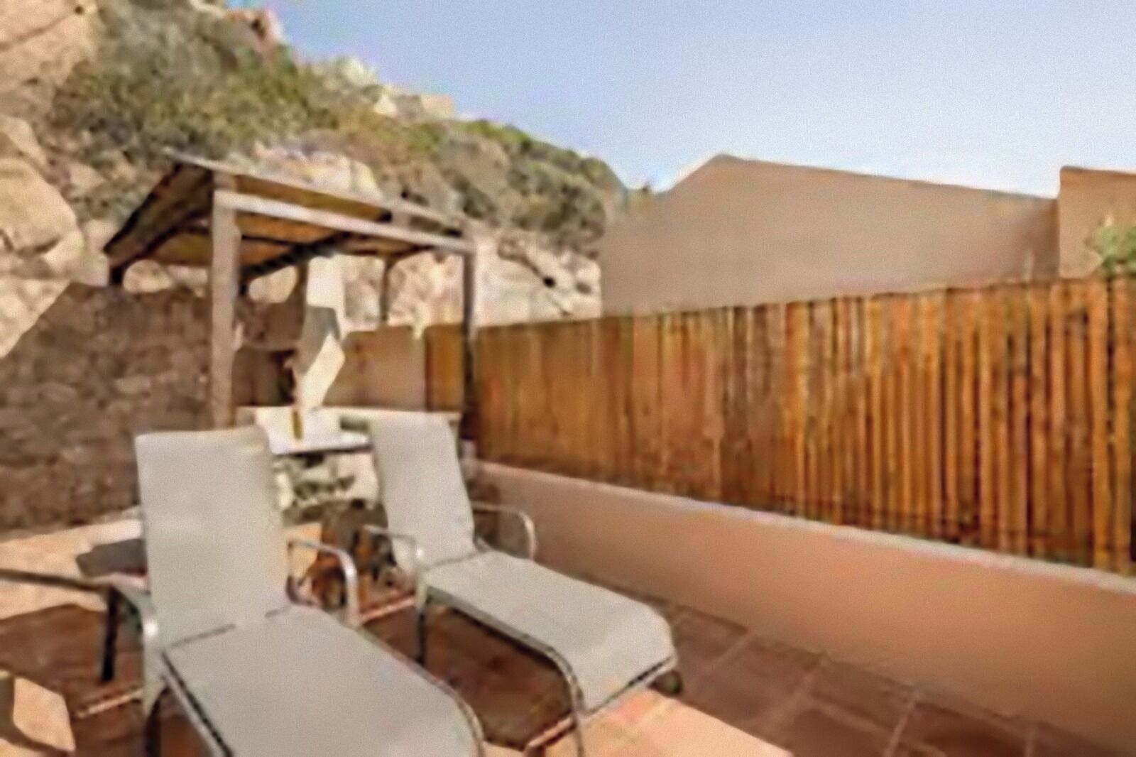 Une terrasse extérieure avec une table et deux chaises, une pergola en bois et un mur en pierre.
