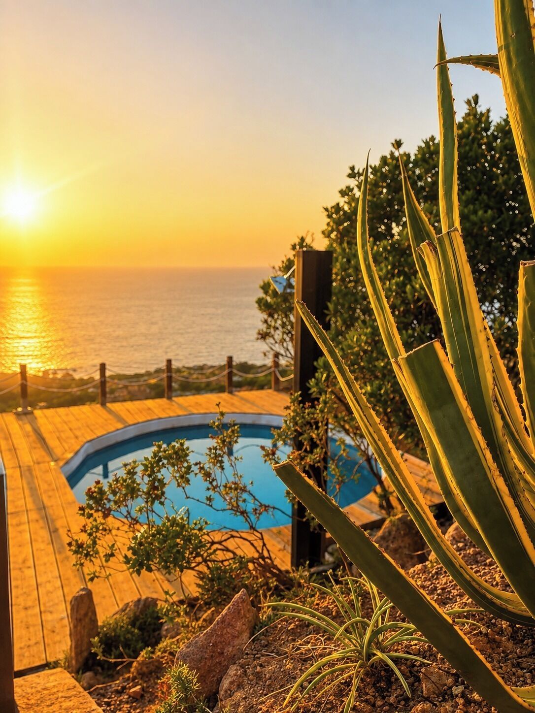 En pool med træterrasse, omgivet af vegetation og med udsigt over vandet i solnedgang.
