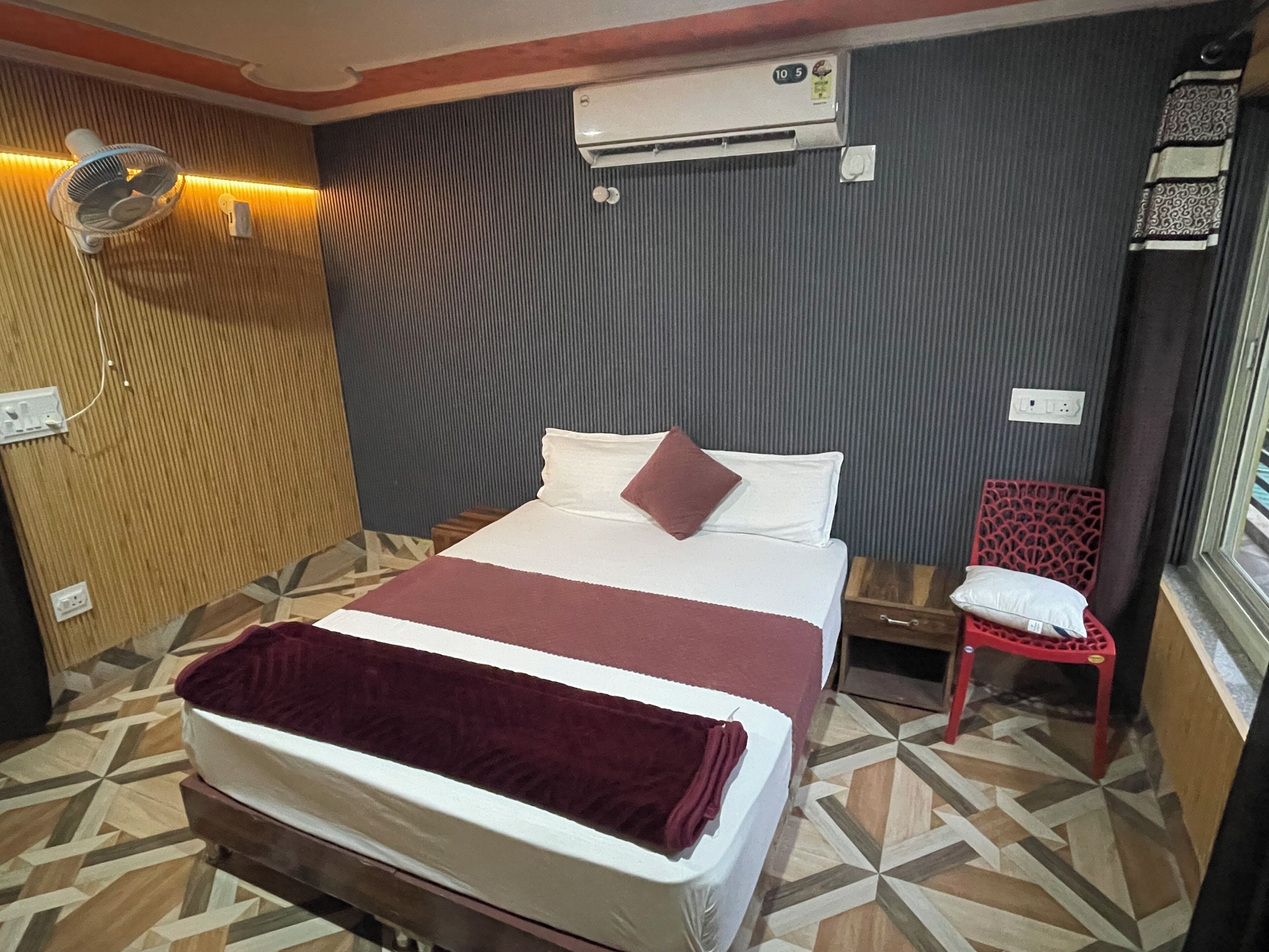 A hotel room with a bed, a bedside table, a red chair, an air conditioning unit, and a wall-mounted fan.