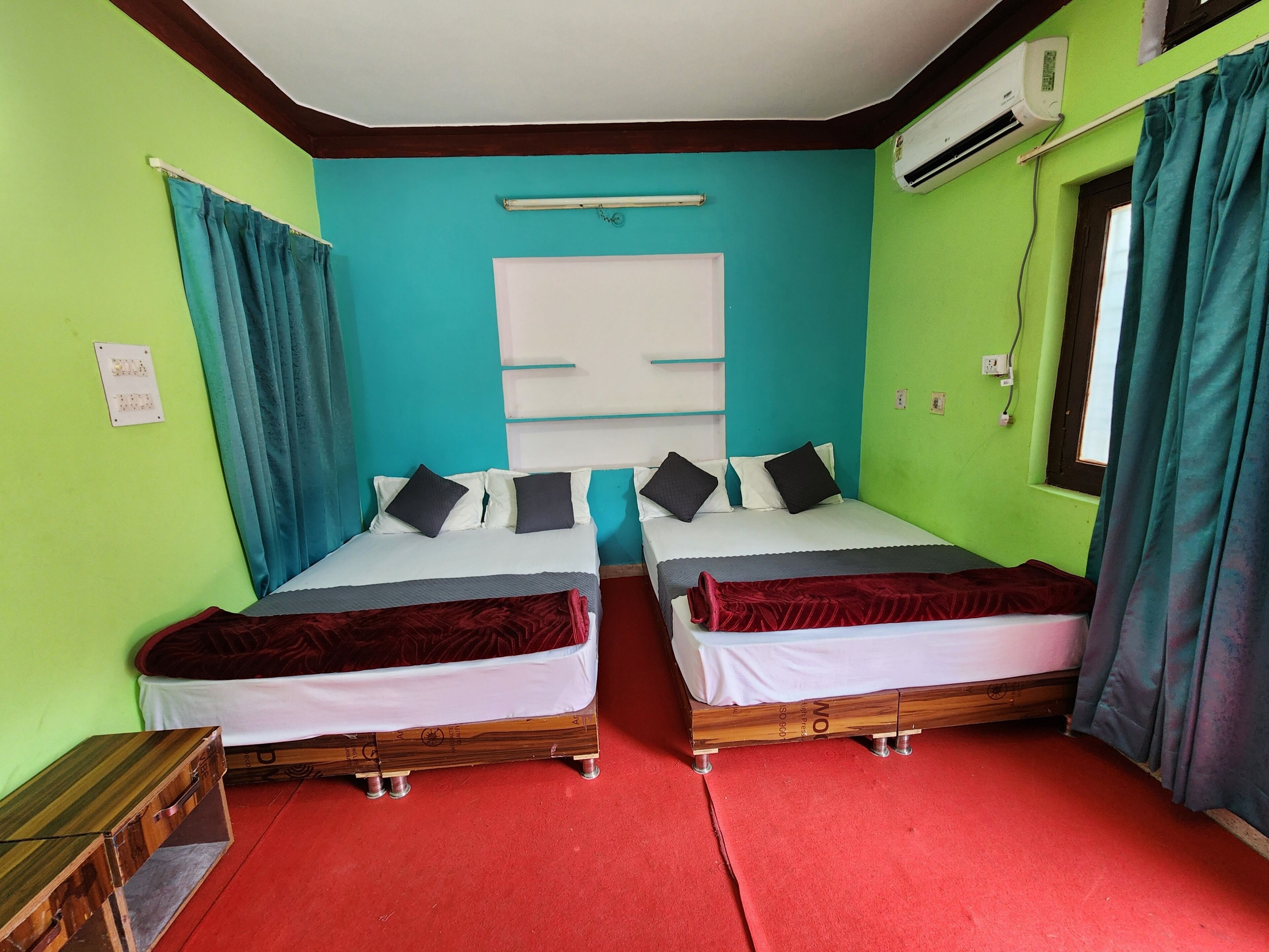 A room with two beds, a window with curtains, an air conditioning unit, and a wooden nightstand.