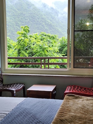 Una habitación con una cama, una pequeña mesa de madera, una silla y una ventana con vistas a un bosque verde exuberante.