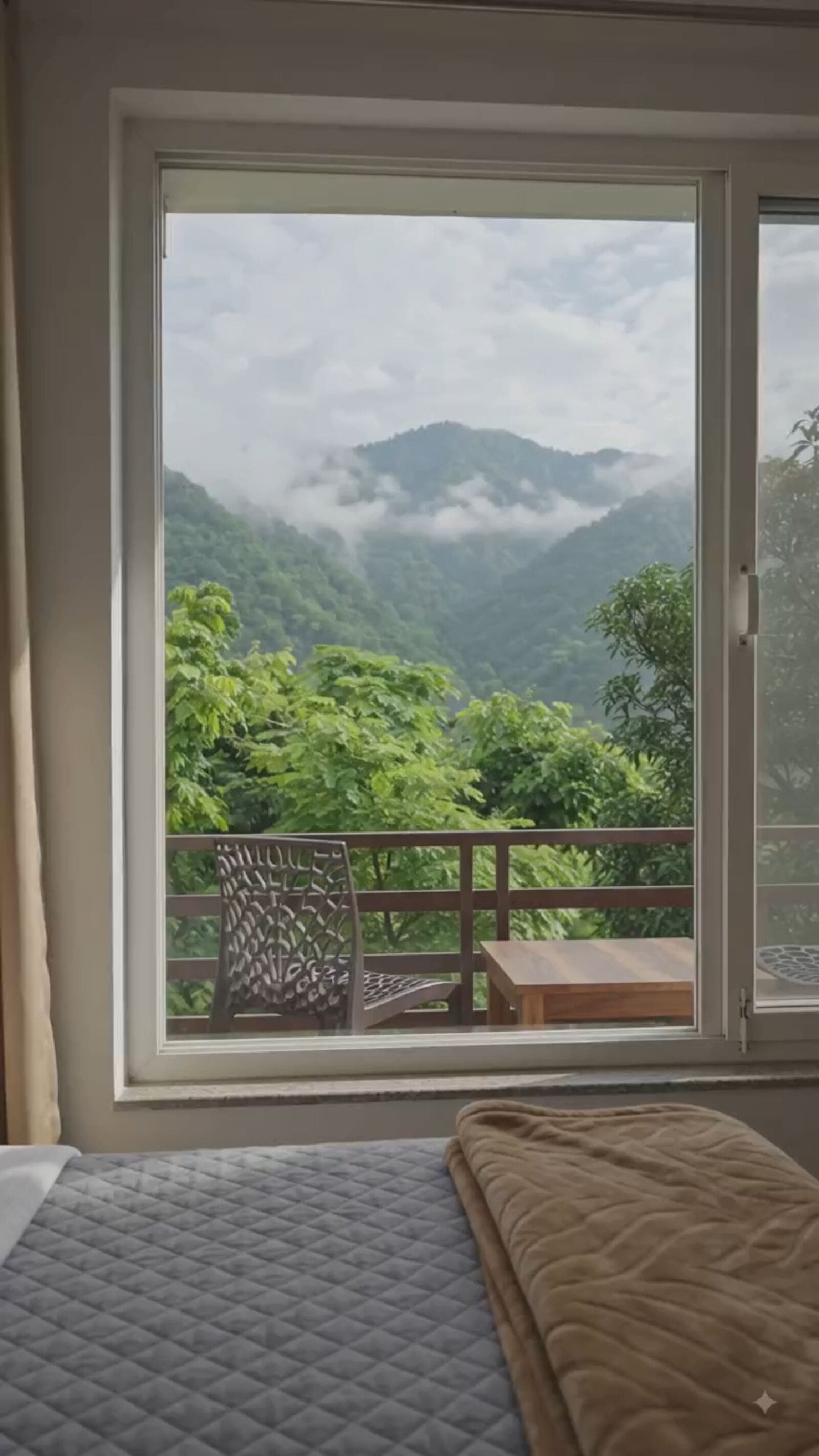 A room with a bed, a window with a view of a mountainous landscape, and a balcony with a chair and table.