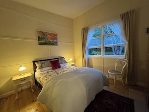 A bedroom with a bed, two bedside tables with lamps, a chair, a window with curtains, and a painting on the wall. - Apple Tree Cottage, a charming mid century hideaway close to town and bushwalks (Blackheath)