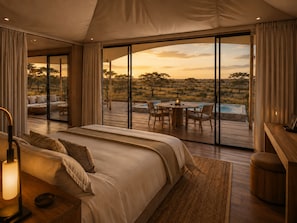 A bedroom with a large bed, a desk, and a view of an outdoor deck with a pool and natural landscape. - Zuraya Serengeti (Makoma)