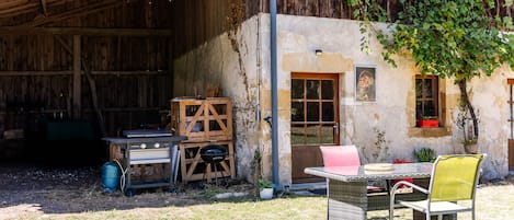 A rustic outdoor area with a wooden table and chairs, a barbecue grill, and a covered storage space.