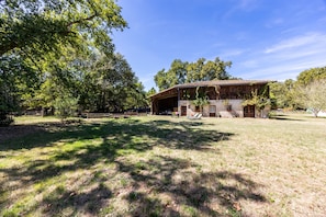 Un bâtiment rustique avec un toit en tuiles, entouré d’une zone herbeuse et d’arbres.