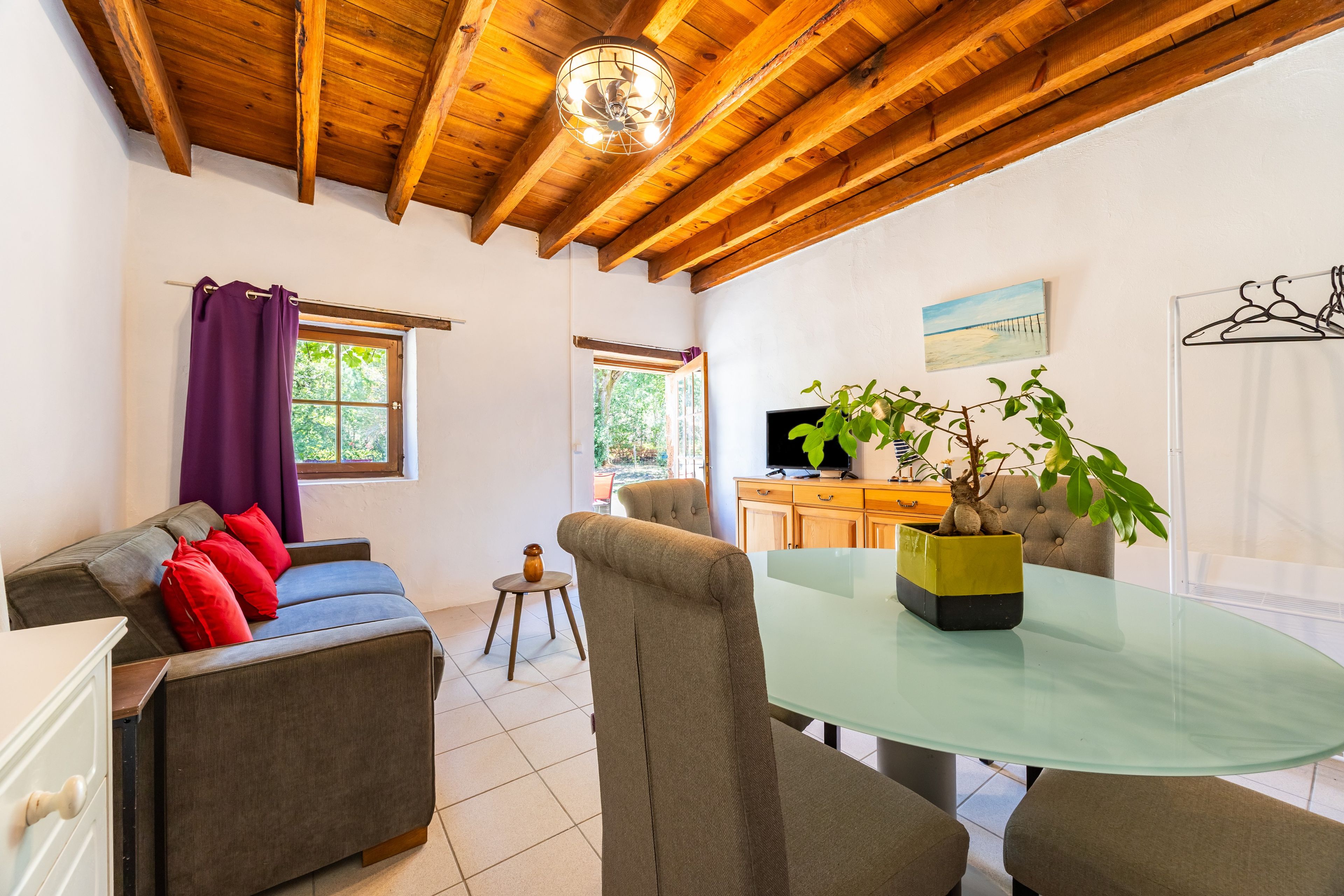 Une pièce avec un plafond en bois, un canapé avec des coussins rouges, une table en verre et une plante dans un pot jaune.