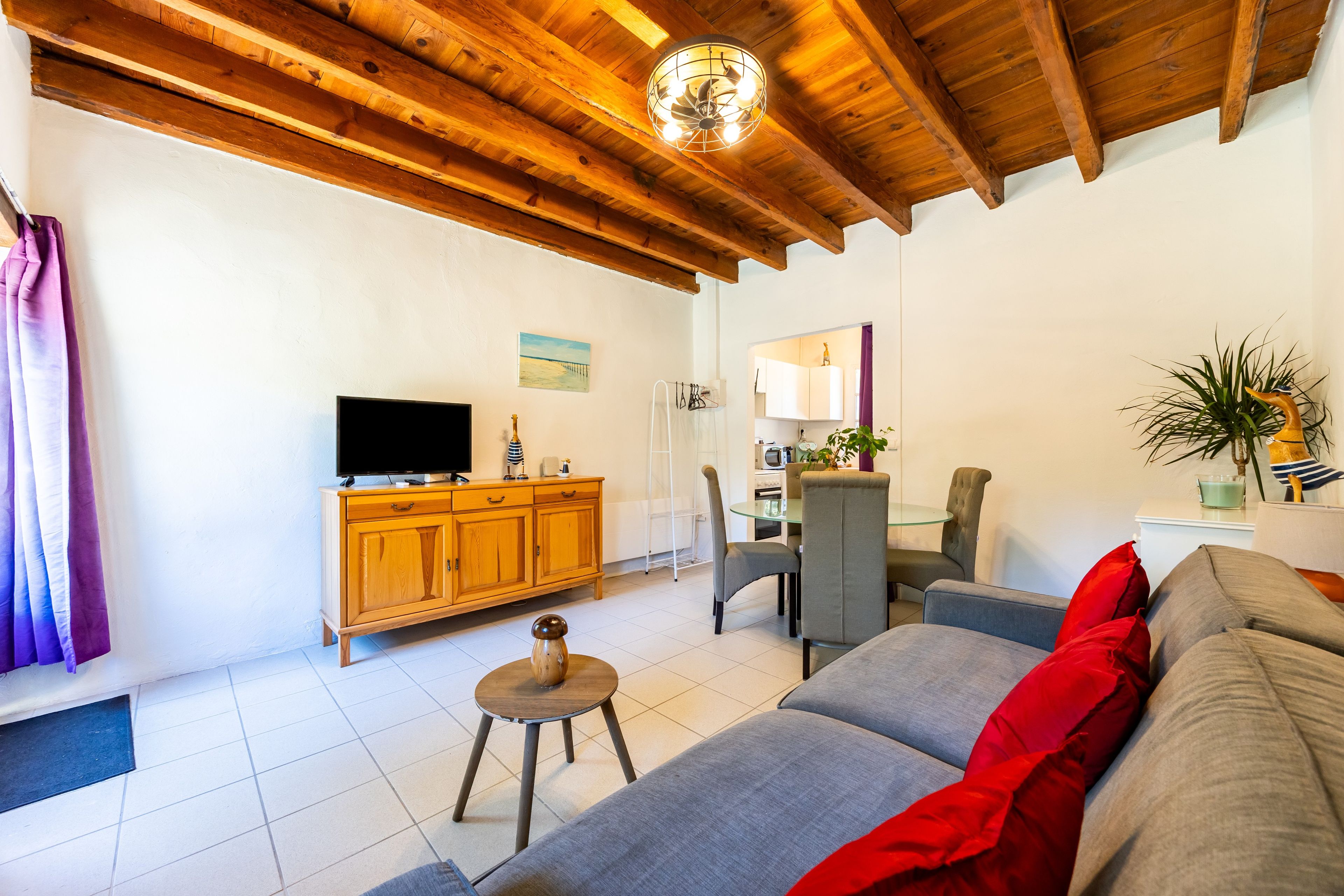 Ein Wohnzimmer mit Holzboden, einem Fernseher auf einer Holzkommode, einem kleinen Hocker, einer Couch mit roten Kissen und einem Essbereich mit Tisch und Stühlen.