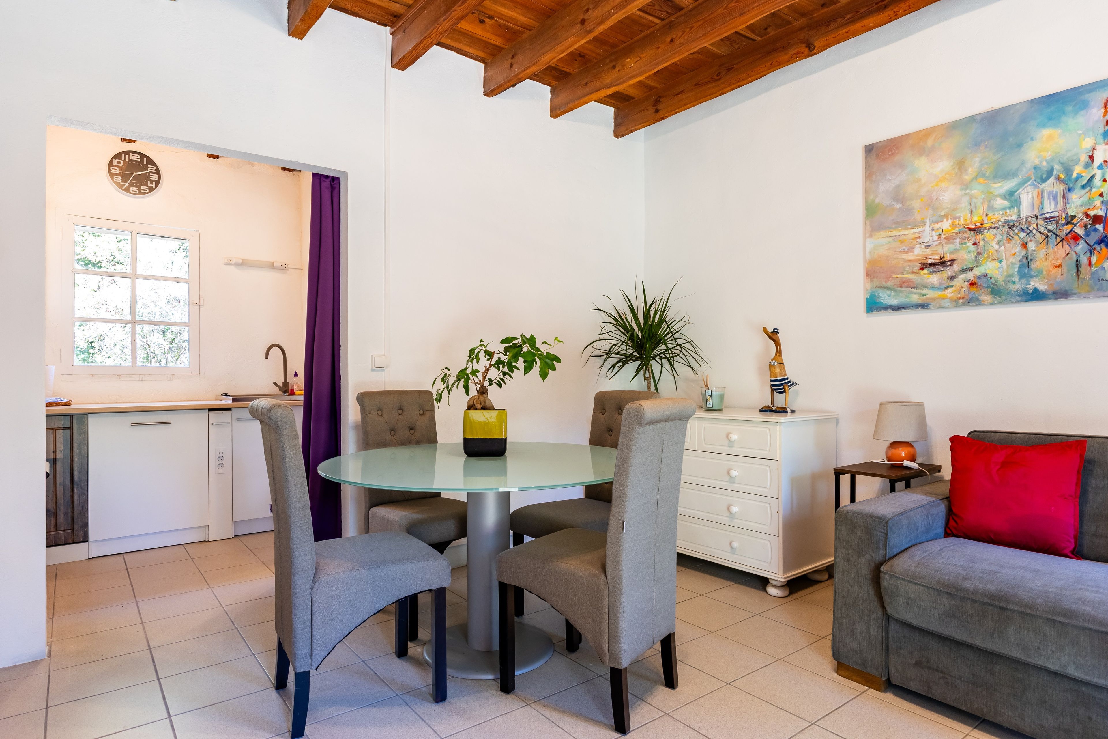 Un salon moderne avec une table à manger au plateau en verre, des chaises à revêtement gris, un canapé gris, une commode blanche et un tableau accroché au mur.