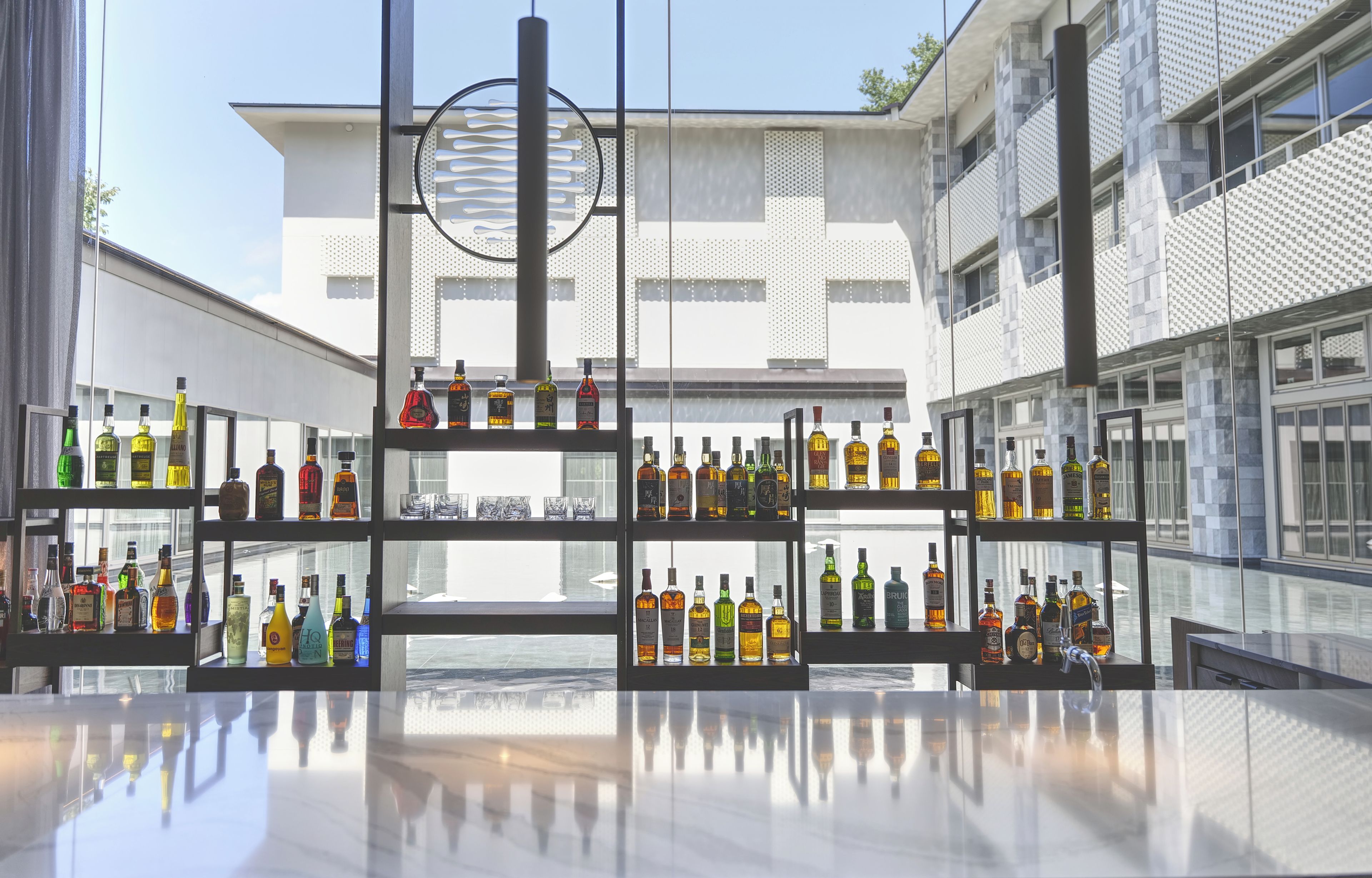Un espace bar moderne, avec de nombreuses étagères remplies de bouteilles d’alcool et de verres, de grandes fenêtres et un bâtiment contemporain en arrière-plan.