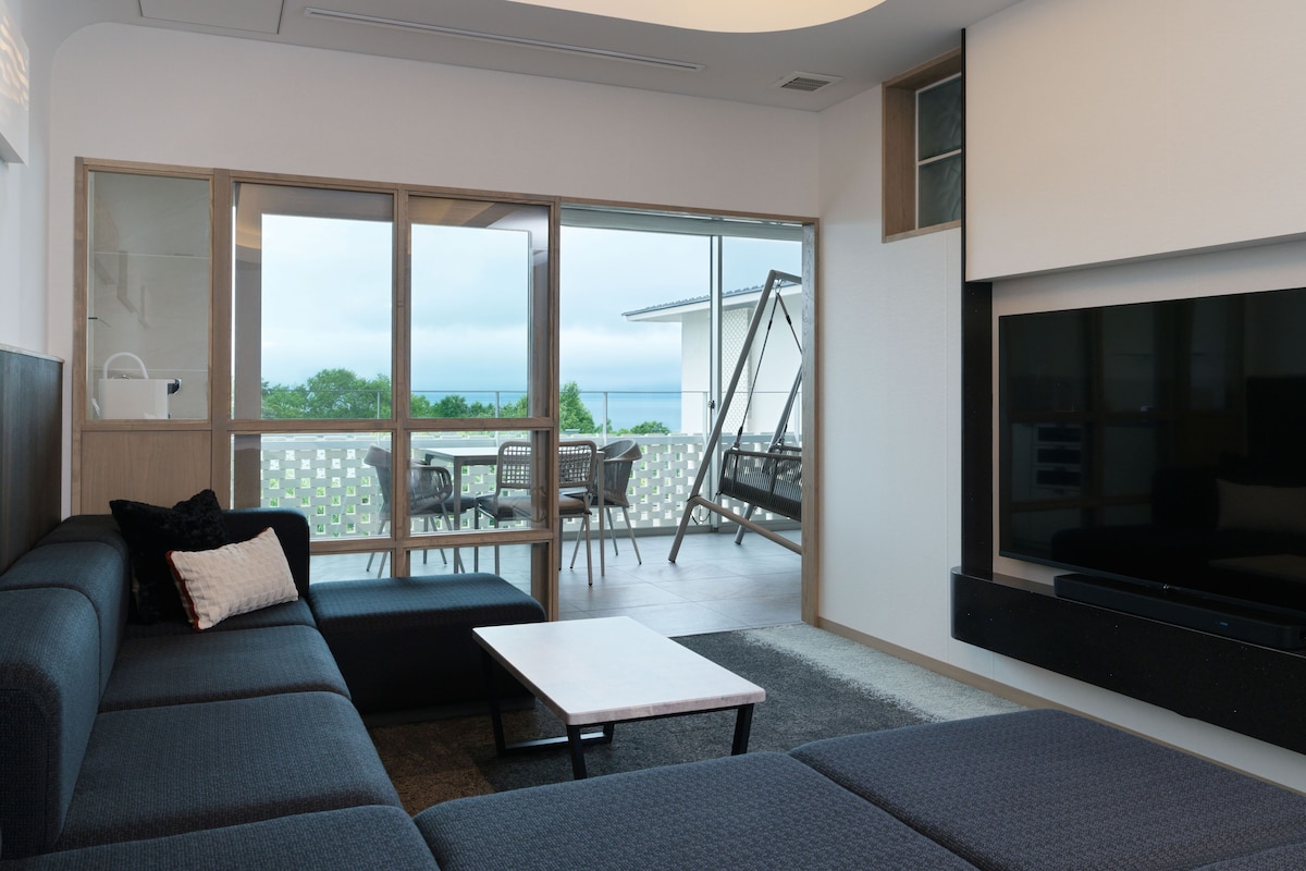 Uma sala de estar moderna com sofá, TV de tela plana e varanda com cadeira de balanço.
